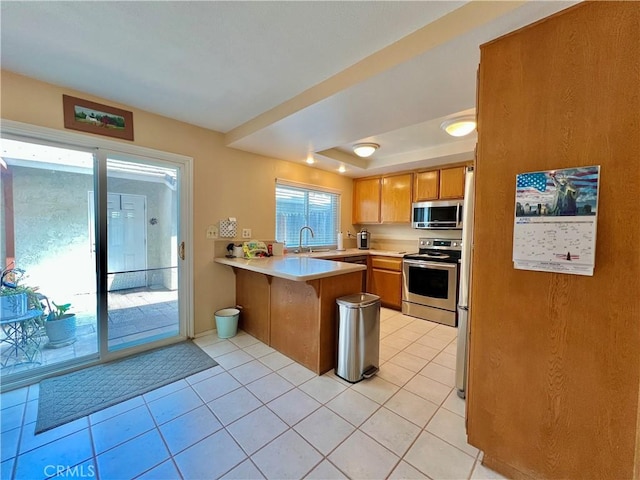 kitchen featuring a peninsula, light tile patterned flooring, light countertops, brown cabinets, and appliances with stainless steel finishes