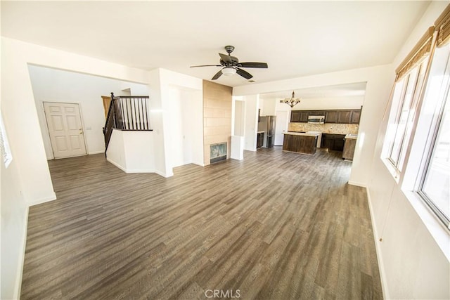 unfurnished living room featuring stairway, dark wood-style floors, ceiling fan with notable chandelier, and baseboards