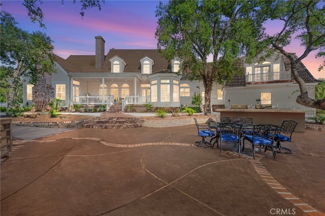 back of property at dusk featuring a chimney, a porch, a balcony, and french doors