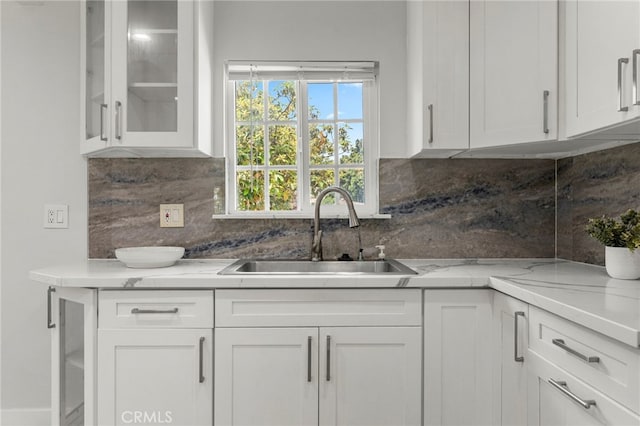 kitchen featuring backsplash, a sink, light stone countertops, glass insert cabinets, and white cabinetry