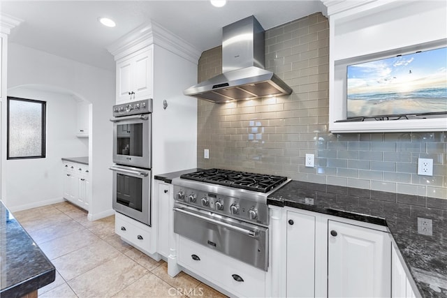 kitchen with double oven, wall chimney exhaust hood, tasteful backsplash, arched walkways, and a warming drawer