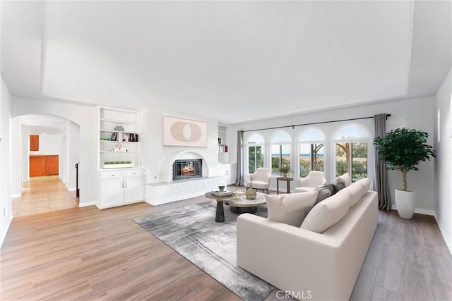 living room featuring built in features, a fireplace, baseboards, light wood-type flooring, and arched walkways