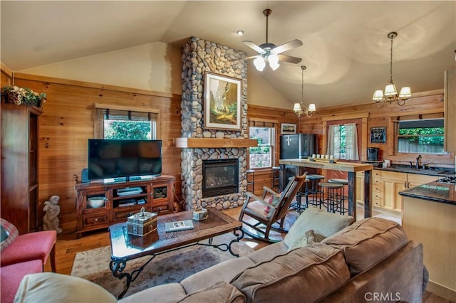living area featuring wood walls, high vaulted ceiling, ceiling fan with notable chandelier, light wood finished floors, and a stone fireplace