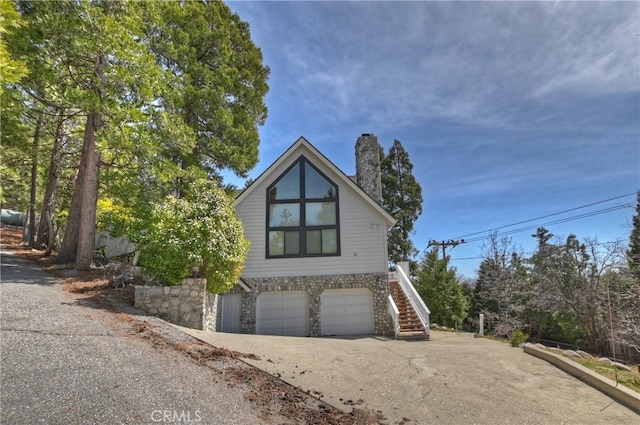rustic home featuring stairway, an attached garage, a chimney, concrete driveway, and stone siding