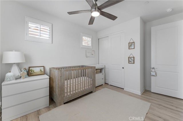 bedroom with light wood-type flooring, baseboards, a closet, ceiling fan, and a crib