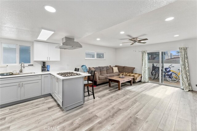 kitchen featuring stainless steel gas cooktop, light countertops, light wood-style flooring, wall chimney exhaust hood, and a sink