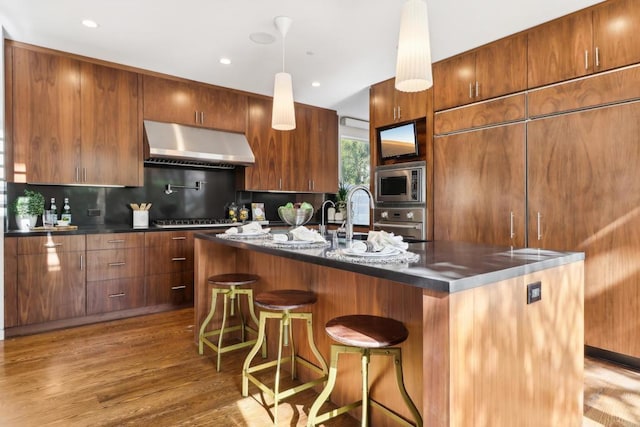 kitchen featuring exhaust hood, appliances with stainless steel finishes, wood finished floors, tasteful backsplash, and a center island with sink