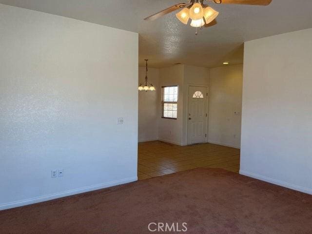 carpeted spare room featuring tile patterned flooring, ceiling fan with notable chandelier, and baseboards