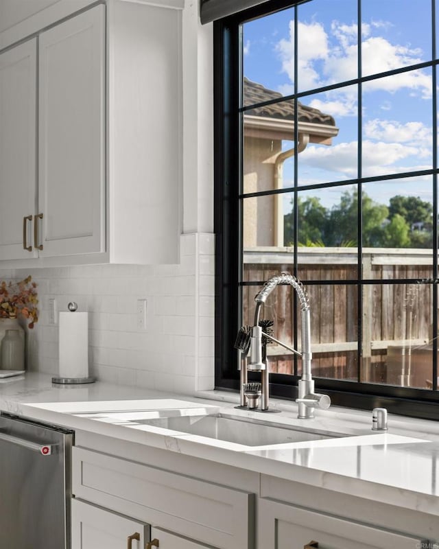 kitchen featuring light stone counters, a sink, backsplash, stainless steel dishwasher, and white cabinets