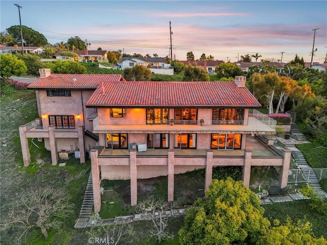 back of house at dusk with a tile roof, a balcony, stairs, a chimney, and stucco siding