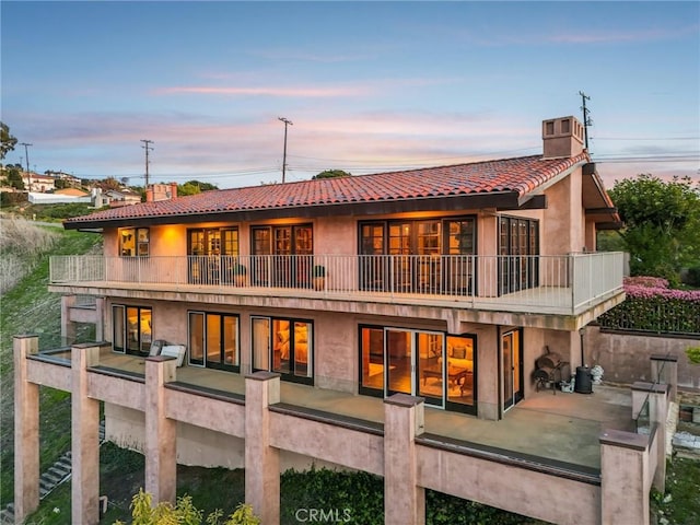 back of property at dusk featuring a chimney, stucco siding, a tiled roof, a patio, and a balcony