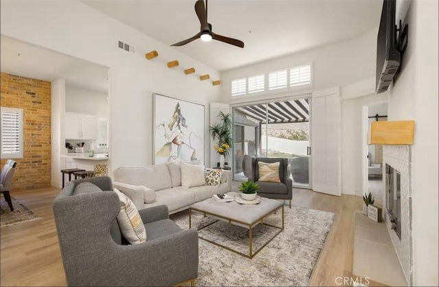 living room featuring visible vents, a towering ceiling, a tiled fireplace, a ceiling fan, and light wood-style floors