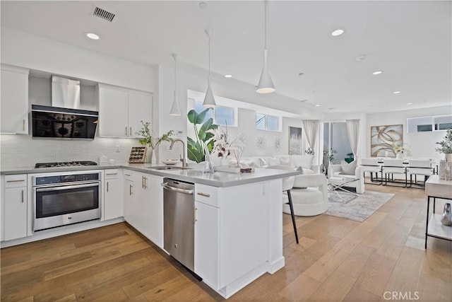 kitchen with visible vents, a peninsula, wall chimney exhaust hood, stainless steel appliances, and a sink