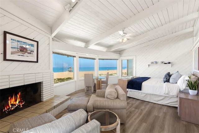 bedroom featuring a warm lit fireplace, beam ceiling, multiple windows, and wood finished floors
