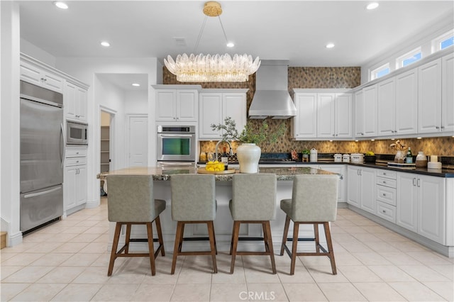 kitchen featuring wall chimney exhaust hood, built in appliances, backsplash, and a breakfast bar area