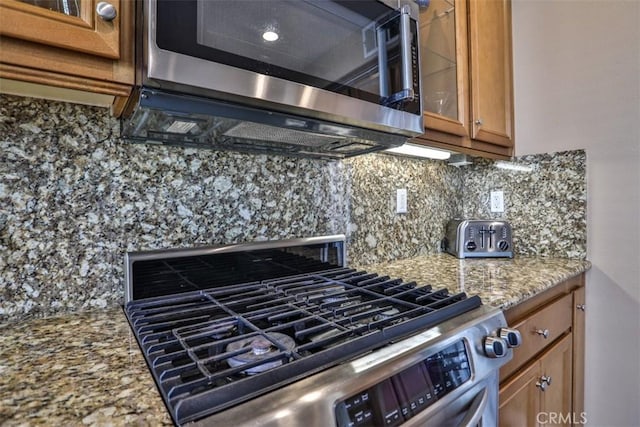 kitchen with stainless steel appliances, decorative backsplash, light stone counters, brown cabinetry, and glass insert cabinets