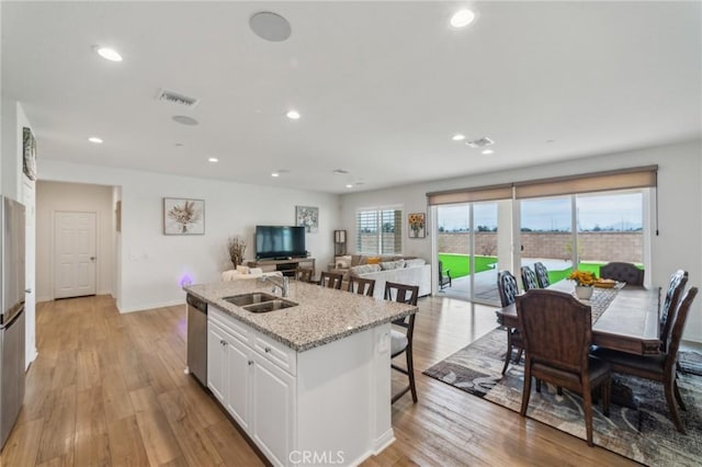 kitchen featuring stainless steel dishwasher, white cabinetry, a kitchen island with sink, a sink, and light wood finished floors