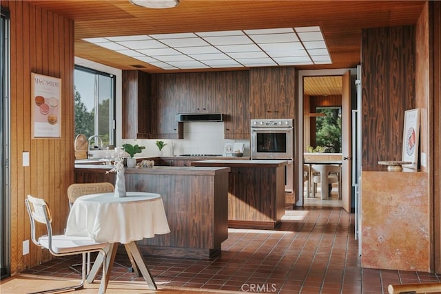 kitchen with decorative backsplash, wooden walls, a peninsula, double oven, and ventilation hood