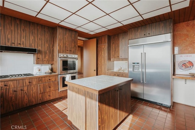 kitchen featuring decorative backsplash, tile counters, stainless steel appliances, under cabinet range hood, and a center island