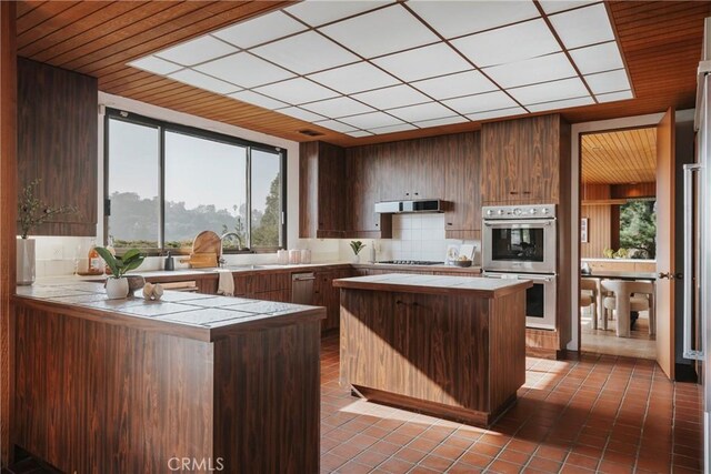 kitchen with a peninsula, under cabinet range hood, tile countertops, double oven, and tile patterned flooring