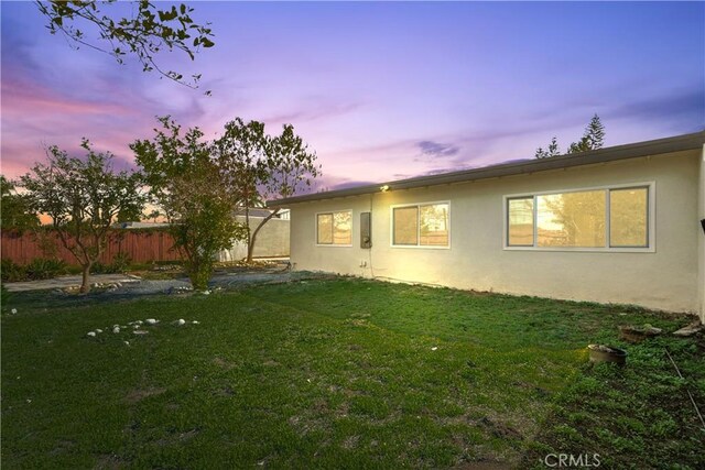 back of house at dusk with stucco siding, fence, and a yard