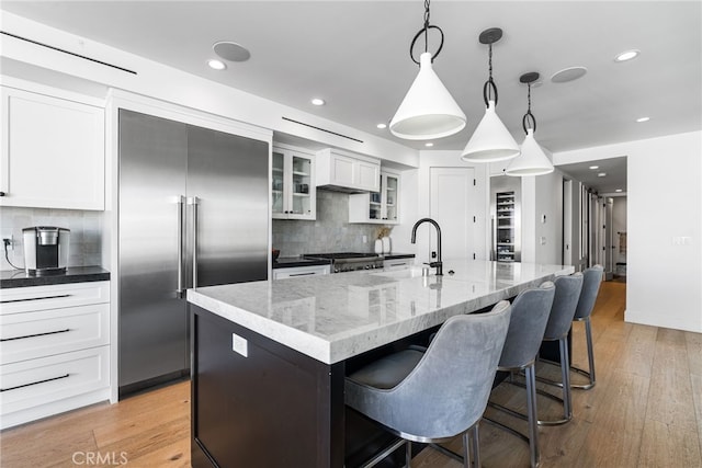 kitchen featuring built in refrigerator, a sink, decorative backsplash, a center island with sink, and white cabinets