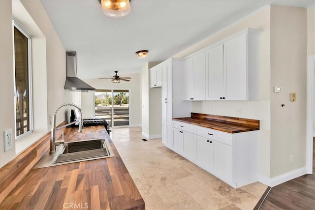 kitchen featuring white cabinets, wooden counters, ceiling fan, wall chimney exhaust hood, and a sink