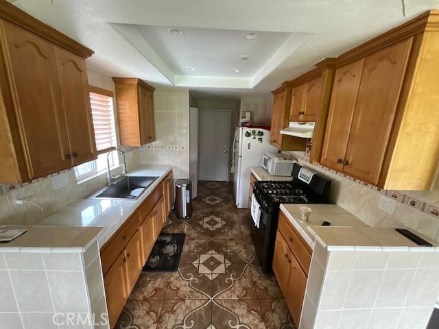 kitchen with backsplash, a sink, a raised ceiling, under cabinet range hood, and white appliances
