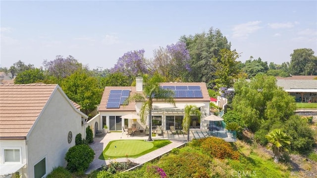 back of house featuring a patio, a chimney, roof mounted solar panels, and stucco siding