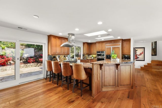 kitchen featuring stainless steel double oven, light stone counters, island range hood, and brown cabinets