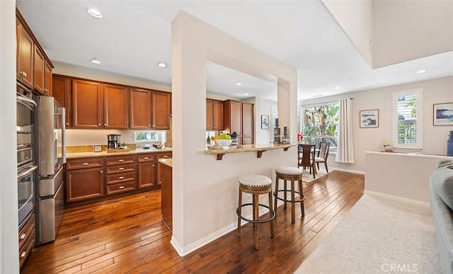 kitchen with brown cabinetry, open floor plan, a breakfast bar area, dark wood-type flooring, and stainless steel appliances