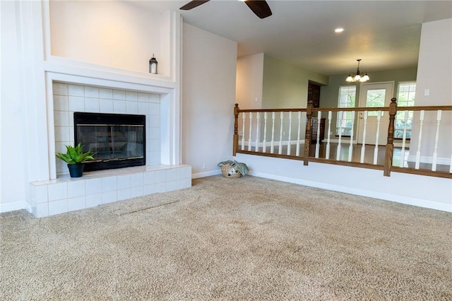 unfurnished living room with baseboards, a tiled fireplace, carpet, and ceiling fan with notable chandelier