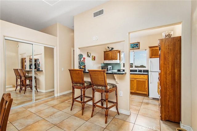 kitchen featuring freestanding refrigerator, dark countertops, light tile patterned flooring, under cabinet range hood, and a breakfast bar area