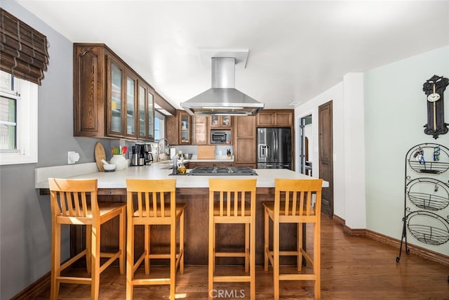 kitchen with glass insert cabinets, wall chimney exhaust hood, appliances with stainless steel finishes, a peninsula, and dark wood finished floors