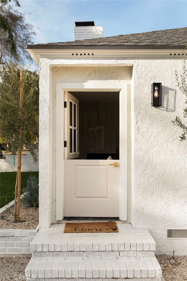 view of exterior entry with a chimney, a shingled roof, and stucco siding