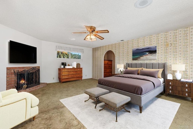 carpeted bedroom featuring arched walkways, a ceiling fan, a textured ceiling, a fireplace, and wallpapered walls