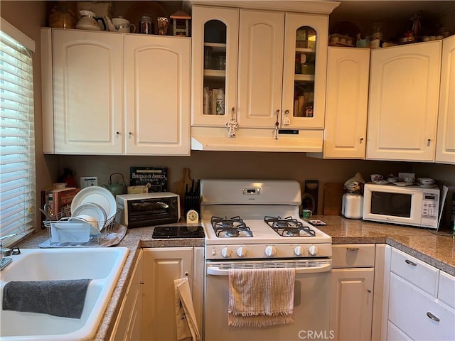 kitchen featuring white cabinetry, white appliances, under cabinet range hood, a sink, and glass insert cabinets