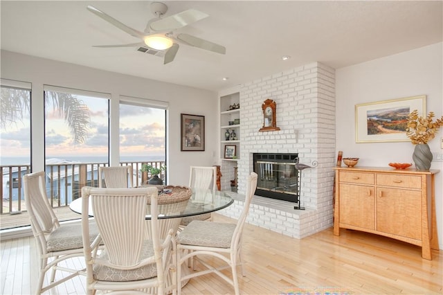 dining room with built in shelves, a ceiling fan, a brick fireplace, and light wood finished floors