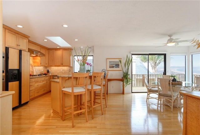 kitchen featuring light wood-type flooring, backsplash, fridge with ice dispenser, under cabinet range hood, and a breakfast bar area