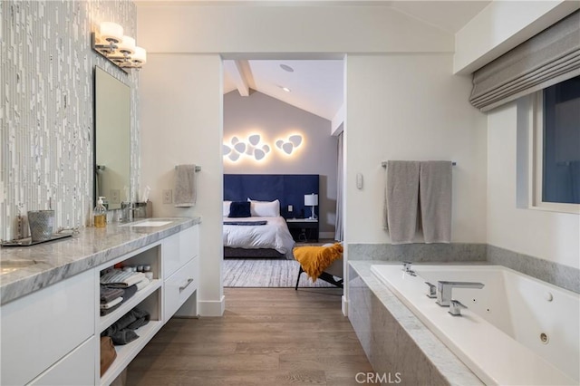 ensuite bathroom with lofted ceiling with beams, connected bathroom, a tub with jets, wood finished floors, and a sink