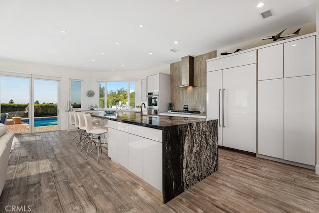 kitchen featuring wall chimney range hood, wood finished floors, a center island with sink, dark stone countertops, and modern cabinets