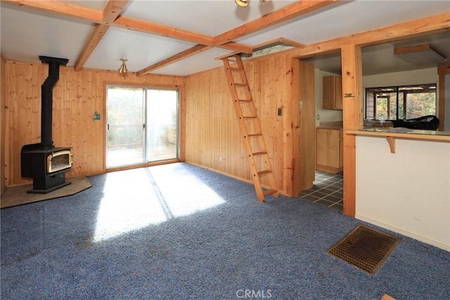 unfurnished living room with wood walls, a wood stove, carpet floors, beam ceiling, and visible vents