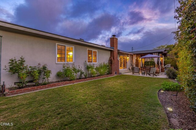 1009 Grandview Ave, Ojai, CA 93023 - back of house at dusk featuring stucco siding, a chimney, a patio area, and a lawn
