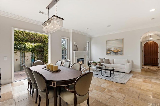 dining space with crown molding, arched walkways, a glass covered fireplace, recessed lighting, and stone tile flooring