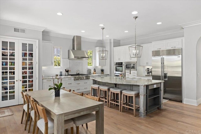 kitchen featuring built in appliances, wall chimney range hood, a kitchen island with sink, visible vents, and a sink