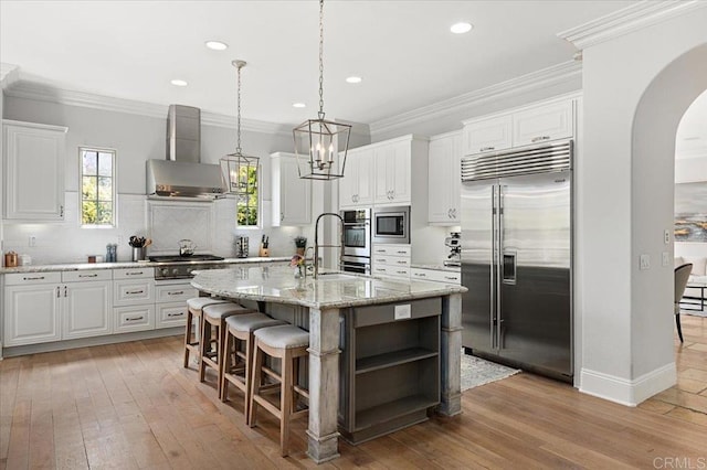 kitchen featuring arched walkways, built in appliances, wall chimney range hood, light wood-style floors, and backsplash