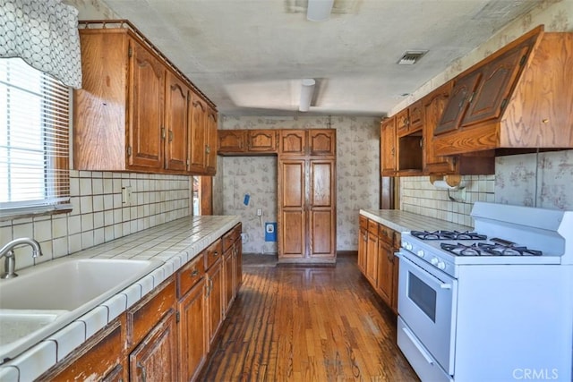 kitchen featuring brown cabinets, white range with gas cooktop, wallpapered walls, dark wood-style flooring, and tile countertops