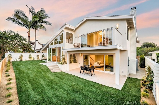 back of property at dusk with a fenced backyard, a chimney, a yard, and a balcony