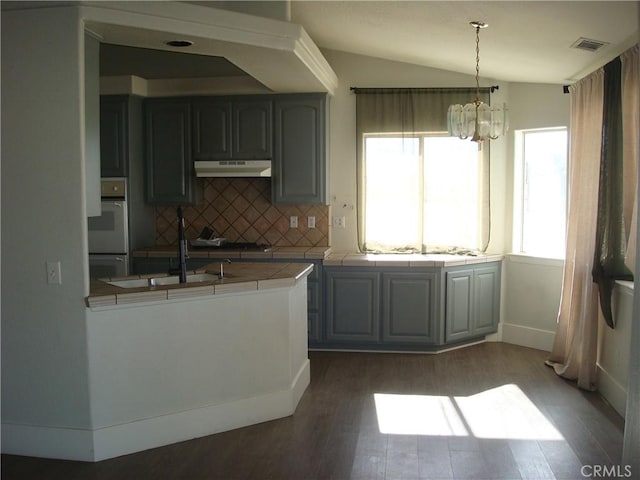 kitchen with tile counters, wood finished floors, tasteful backsplash, under cabinet range hood, and a notable chandelier