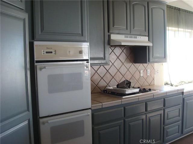 kitchen with backsplash, tile counters, stovetop, white double oven, and under cabinet range hood